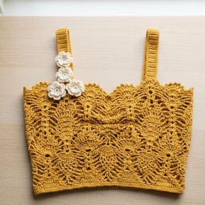 Op de afbeelding: Een mosterdgele gehaakte crop top met een ingewikkeld ananassteekpatroon, een geschulpte halslijn en twee brede schouderbandjes. Drie kleine, gebroken witte gehaakte bloemen zijn bevestigd aan de basis van het linker schouderbandje.
