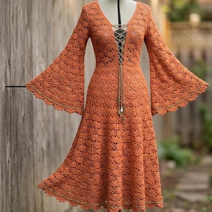 Puede incluir: Un vestido naranja de ganchillo con mangas largas y acampanadas y un escote en V. El vestido presenta un detalle de cordones en el busto y una falda fluida. Las mangas y el dobladillo están adornados con un borde dorado. El vestido se exhibe en un maniquí.