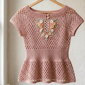 May include: A dusty rose crochet top with short sleeves and a peplum waist. The top features a floral embroidery around the neckline and a button closure. The openwork design adds a delicate touch to this handmade garment.