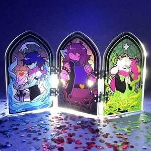 May include: A triptych of acrylic panels featuring stylized character art. Each panel depicts a different character in a stained-glass window design, with vibrant colors and intricate details. The panels are connected and stand upright, with a scattering of confetti in the foreground.