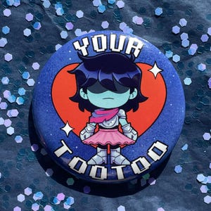 May include: A circular pinback button featuring a cartoon character with dark hair, light blue skin, and silver armor, wearing a pink skirt. The character is centered over a red heart on a dark blue background with the text YOUR TOOTOO.
