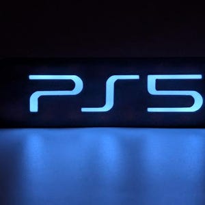 Peut inclure: Une enseigne rectangulaire de couleur sombre présentant le texte PS5 illuminé dans une couleur bleu clair vive et éclatante. Les lettres et le chiffre sont stylisés avec des lignes épurées et modernes, contrastant avec le fond sombre.