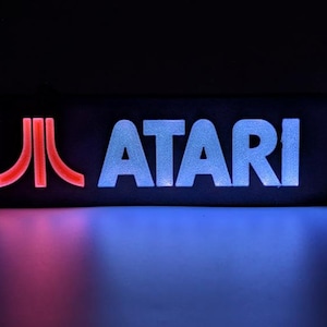 Può includere: Un'insegna rettangolare con il logo rosso Atari a sinistra e la parola ATARI in lettere maiuscole bianche a destra. L'insegna è illuminata su uno sfondo scuro, proiettando un tenue bagliore rosso e blu sulla superficie sottostante.
