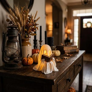 May include: A white, glowing ghost figurine holding a small brown book, positioned on a wooden surface. The ghost has hollow, illuminated eyes and a draped, sheet-like appearance. It is surrounded by small pumpkins, a lantern, and dried wheat.