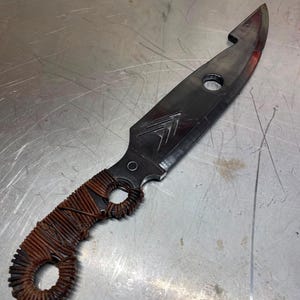 Könnte beinhalten: Ein Messer nach Maß mit einer dunklen, verwitterten Klinge und einem umwickelten Griff. Die Klinge hat eine einzigartige Form mit einem Loch und einer Kerbe in der Nähe der Spitze. Der Griff ist mit braunem Draht umwickelt. Das Messer scheint handgefertigt zu sein.
