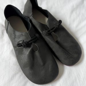 May include: A pair of dark charcoal gray slip-on shoes featuring a rounded toe, a low-cut topline, and a decorative black rope knot closure detail on the upper vamp. The shoes have a matte, textured finish and a simple, unadorned construction.