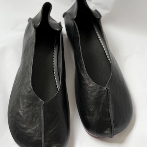 May include: A pair of black leather slip-on shoes featuring a soft, unstructured silhouette with a center seam running down the front, a rounded toe, and visible white stitching along the inner edge of the foot opening.