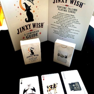 May include: A Jinxy Wish fortune telling playing card set including a companion guide book, two white card boxes, and three individual cards featuring a stork, a cow, and a safe. The text reads Jinxy Wish Fortune Telling Playing Cards.