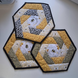 Può includere: Tre tappetini in tessuto trapuntato esagonali con un design patchwork di motivi a nido d'ape gialli, stampe di foglie bianche e nere e centri in tessuto bianco decorati con illustrazioni di bombi e favi dorati, rifiniti con bordi neri.
