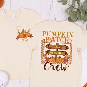 May include: A cream-colored short-sleeve t-shirt featuring a small chest graphic of pumpkins and the name Emily, and a large back graphic with the text PUMPKIN PATCH FALL MEMORIES CREW, wooden signpost illustrations, and orange pumpkins.