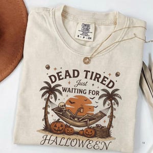 May include: A cream-coloured short-sleeve t-shirt featuring a graphic of a skeleton relaxing in a hammock between two palm trees. The text reads DEAD TIRED JUST WAITING FOR HALLOWEEN. The design includes pumpkins, bats, and a sunset background.