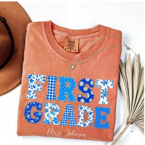 Op de afbeelding: Een terracotta-kleurig t-shirt met de tekst FIRST GRADE in blauwe en witte stoffen letters met patronen. Het shirt heeft een ronde hals en een Comfort Colors label. Onder de letters staat de naam Miss Johnson in witte letters.