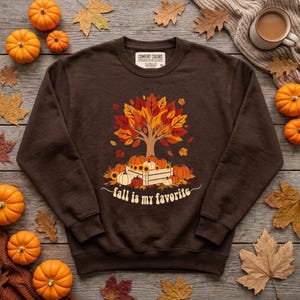 May include: A dark brown crewneck sweatshirt featuring a graphic of a tree with orange, red, and yellow leaves above a wooden crate filled with pumpkins and sunflowers. The text "fall is my favorite" is printed in white below the crate.