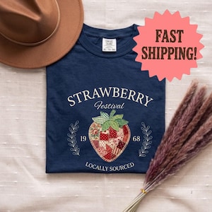 Peut inclure: Un t-shirt bleu marine avec un motif central de fraise en patchwork et des feuilles vertes. Le texte indique STRAWBERRY Festival, 1968, et LOCALLY SOURCED. Un chapeau fedora marron et un bouquet d'herbes de la pampa séchées sont posés à côté.