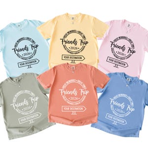 Puede incluir: Seis camisetas de manga corta y cuello redondo en azul claro, amarillo, rosa claro, verde salvia, coral y azul medio. Cada camiseta presenta un gráfico circular con el texto "Friends Trip 2026" y "YOUR DESTINATION" en un icono de señal.