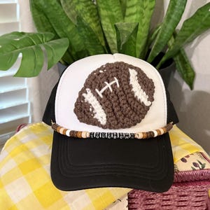 Pode incluir: Boné de caminhoneiro preto e branco com um design de futebol americano de crochê. A bola é marrom com listras brancas. O chapéu tem uma faixa de contas com a palavra "Touchdown".