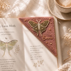 May include: A pink felt bookmark with an embroidered luna moth design and the name "Emma". The bookmark is placed on an open book with text about the luna moth and its symbolism. A cup of coffee is in the background.