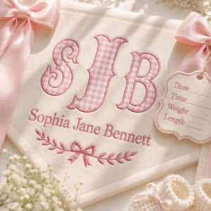 Könnte beinhalten: Ein cremefarbenes Stoffbanner mit rosa kariert gestickten Initialen SJB, dem Namen Sophia Jane Bennett sowie einem Schleifen- und Blattmotiv. Es enthält zwei rosa Satinbänder und ein Anhängeschild mit Feldern für Date, Time, Weight und Length.