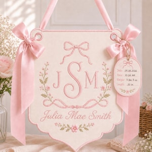 Può includere: Un arazzo da parete in tessuto color crema con ricami rosa che presenta il monogramma JSM, il nome Julia Mae Smith e motivi floreali. Include fiocchi in raso rosa e un'etichetta pendente con il testo: Date: 05.08.2026, Time: 10:24 AM, Weight: 7 lbs 8 oz, Length: 20 in.