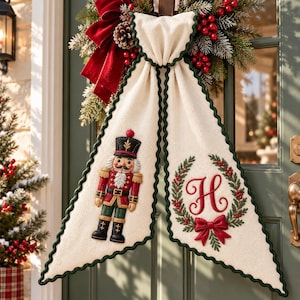 May include: A cream-colored fabric door hanger featuring a dark green scalloped trim. The left panel displays an embroidered nutcracker in red, green, and gold. The right panel features an embroidered red letter H inside a green wreath with a red bow.