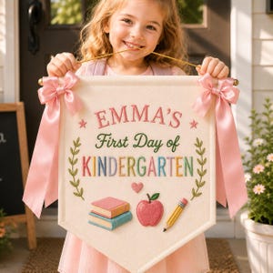 May include: A cream-colored fabric banner featuring the embroidered text "EMMA'S First Day of KINDERGARTEN" in pink, green, and multicolored letters. The banner is decorated with two pink satin bows, a stack of books, an apple, and a pencil.