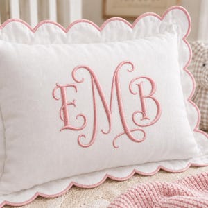 May include: A rectangular white pillow featuring a scalloped edge trimmed in pink thread. The center of the pillow displays a large, embroidered monogram in pink thread with the letters E, M, and B in a decorative, cursive script font.