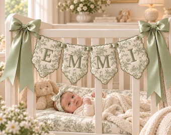 May include: A white crib featuring a decorative fabric bunting with the name EMMI embroidered in green. The bunting is adorned with a green toile pattern and is attached to the crib with large, sage green satin bows on each side of the frame.