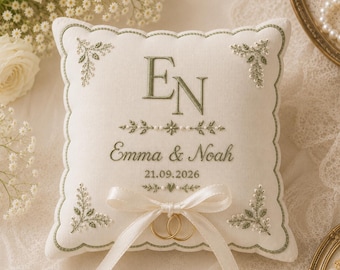 May include: A square off-white ring bearer pillow featuring sage green embroidery of the initials EN, the names Emma & Noah, and the date 21.09.2026. The pillow is decorated with floral corner motifs and a cream satin ribbon holding two gold rings.