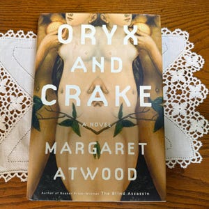 May include: A paperback book titled Oryx and Crake, a novel by Margaret Atwood. The cover features a symmetrical, mirrored illustration of human figures with pale skin, green leaves, and white text. The author is noted as the author of The Blind Assassin.