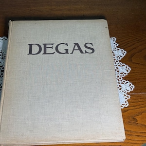 Könnte beinhalten: Ein Hardcover-Buch mit einem hellbeigen Stoffeinband, auf dessen Vorderseite das Wort DEGAS in dunkelbraunen Großbuchstaben aufgedruckt ist. Das Buch ist rechteckig, hat ein schlichtes Design und einen sichtbaren Buchrücken an der linken Seite.