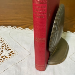 May include: A red hardcover book with gold lettering on the spine, titled 'Catriona' by Robert Louis Stevenson, held upright by a brass-colored metal bookend shaped like a seashell. The book spine features decorative gold geometric patterns.