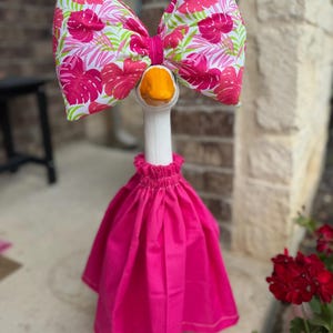 May include: A decorative goose figurine wearing a bright pink skirt with an elastic waistband and a large matching bow on its head. The bow features a vibrant tropical pattern of pink monstera leaves and green palm fronds on a white background.