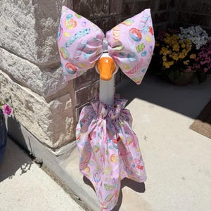 May include: A decorative goose statue wearing a light pink fabric outfit featuring a colorful candy and donut print. The ensemble includes a large, structured bow headpiece and a ruffled, gathered skirt that drapes around the goose's neck and body.