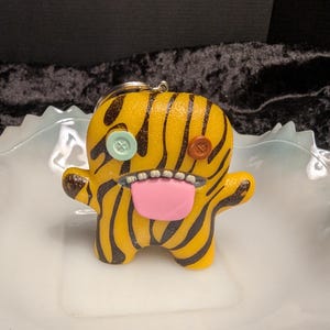 May include: A yellow keyring with black tiger-like stripes, featuring a light blue button eye on the left and a brown button eye on the right. The figure has a wide, open mouth with visible white teeth and a large, protruding pink tongue.