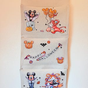 May include: A beige fabric wall hanging organizer with three pockets featuring colorful Halloween-themed illustrations of cartoon characters, pumpkins, and the text "HAPPY HALLOWEEN" in a decorative font, suspended from a wooden dowel rod.
