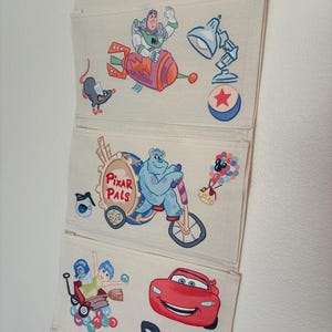 May include: A three-tiered, off-white fabric organizer featuring colorful cartoon character illustrations. The top section shows Buzz Lightyear on a rocket, the middle features Sully on a tricycle, and the bottom has Lightning McQueen. A fun and functional storage solution.