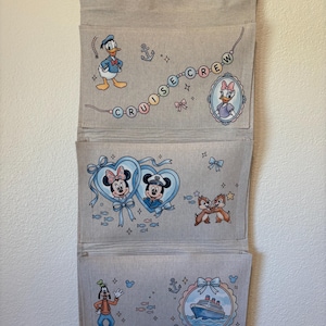 May include: A beige fabric wall organizer with three pockets, featuring Disney characters. Donald Duck, Daisy Duck, Mickey and Minnie Mouse, Chip and Dale, and Goofy are depicted. The text "CRUISE CREW" is also present, along with anchors and a cruise ship illustration.