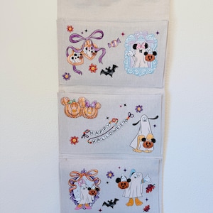 May include: A beige fabric wall hanging organizer with three pockets featuring colorful Halloween-themed illustrations of cartoon characters in costumes, pumpkins, bats, flowers, and the text "HAPPY HALLOWEEN" printed in a decorative font.