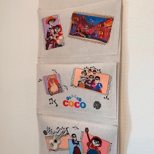 May include: A beige fabric wall hanging organizer with three pockets featuring colorful printed scenes and characters from the movie Coco. The design includes illustrations of Miguel, Hector, and other characters, musical notes, a guitar, and the Coco logo.