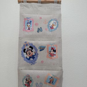 May include: A beige fabric wall hanging organizer with three pockets, featuring printed illustrations of Disney characters including Daisy Duck, Donald Duck, Mickey Mouse, Minnie Mouse, Goofy, and Pluto framed in pastel colors with nautical motifs.