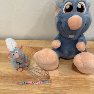 May include: A large blue plush rat wearing a white chef hat, a smaller blue plush rat wearing a white chef hat holding a metal wire whisk, and a beaded bracelet with grey and pink beads featuring the name REMY in white lettered beads.