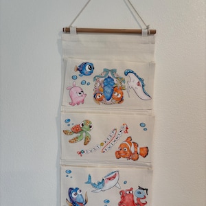 May include: A white fabric wall organizer with four pockets, each featuring colorful illustrations of characters from the movie "Finding Nemo." The top pocket has a blue fish, a pink fish, and other characters. The third pocket has a turtle, a clownfish, and the text "Just Keep Swimming."