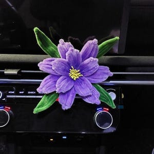 Pode incluir: Um clipe de flor roxa para saída de ar de carro com uma superfície texturizada e felpuda. A flor apresenta várias pétalas roxas em camadas, um centro amarelo brilhante e várias folhas verdes que se estendem por trás das pétalas.
