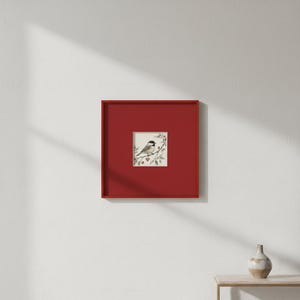 May include: A framed print featuring a small bird perched on a branch with red berries. The artwork is set within a square, deep red frame, mounted on a white wall. The print's colors include shades of gray, white, and red.