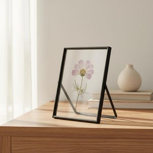 Op de afbeelding: Een rechthoekige zwevende fotolijst met een dunne zwarte metalen rand. De lijst bevat een enkele geperste lichtroze cosmosbloem met een geel hart en groene steel, weergegeven tussen twee heldere glasplaten met een metalen standaard.