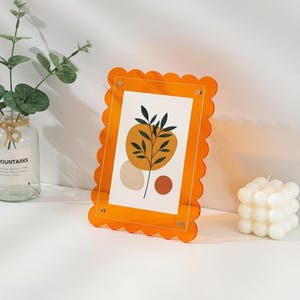 Puede incluir: Un marco de fotos rectangular con un borde festoneado de color naranja brillante hecho de acrílico. El marco contiene una impresión blanca con una ilustración botánica central de hojas verde oscuro, un círculo grande amarillo mostaza, un óvalo beige y un círculo pequeño color óxido.