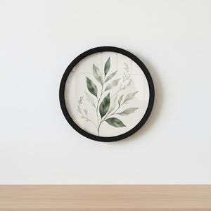 May include: A circular wall art print with a watercolour illustration of green and grey leaves and stems. The artwork is framed in a black circular frame, creating a minimalist and natural aesthetic. The art piece is displayed against a white background.