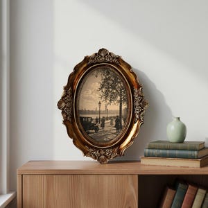 May include: An oval, ornate gold picture frame with a sepia-toned print of a vintage street scene. The frame has a detailed, scrolled design. The image is displayed on a wooden cabinet with books and a small vase.