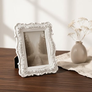 May include: A rectangular white picture frame featuring an ornate, raised relief pattern of roses and small embedded crystals along the border. The frame holds a vertical photograph depicting a path through a forest of tall, dark trees.