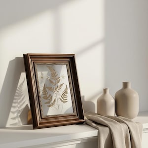May include: A framed botanical print featuring pressed ferns and delicate white flowers. The frame is a rich brown with a beaded border. Two neutral-toned vases and a folded beige cloth are also visible.